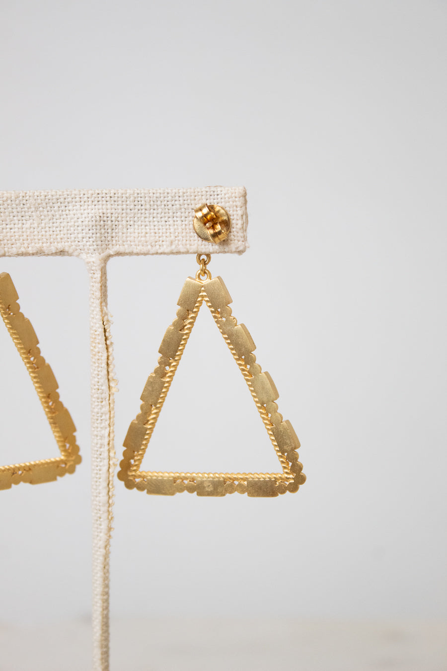 Gold triangular dangle earrings with a textured beaded border, displayed on a beige fabric jewelry stand against a light gray background.