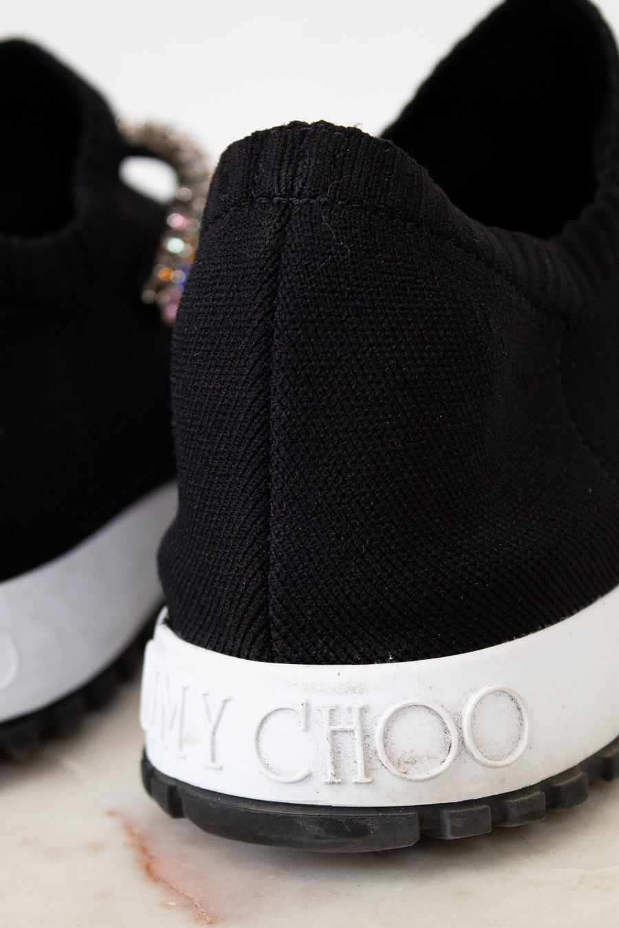 Black sneaker with white sole featuring 'Jimmy Choo' branding on a marble surface.