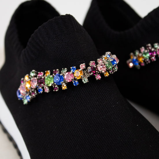 Black slip-on shoes with colorful gemstone embellishments on a light gray background