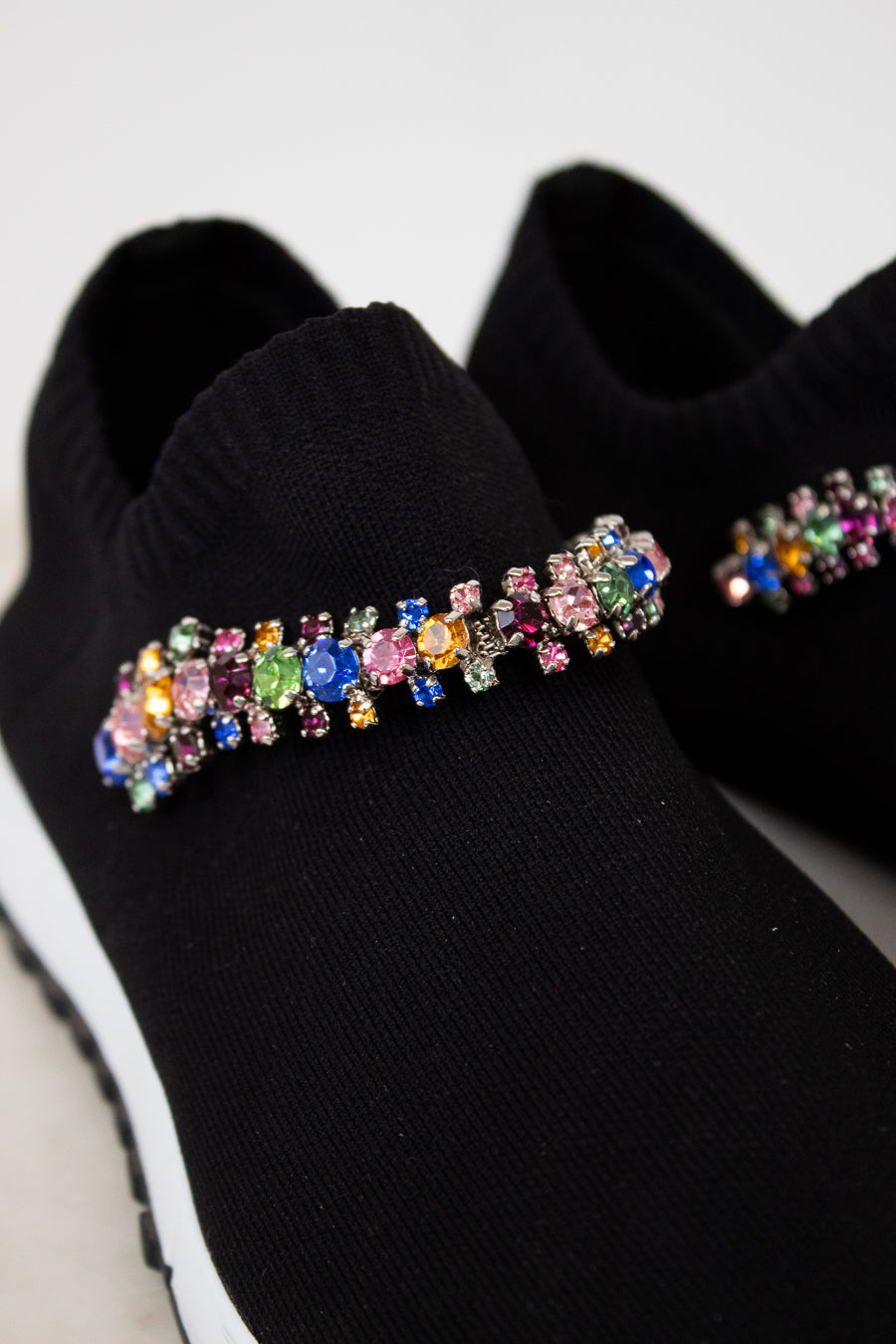 Black slip-on shoes with colorful gemstone embellishments on a light gray background
