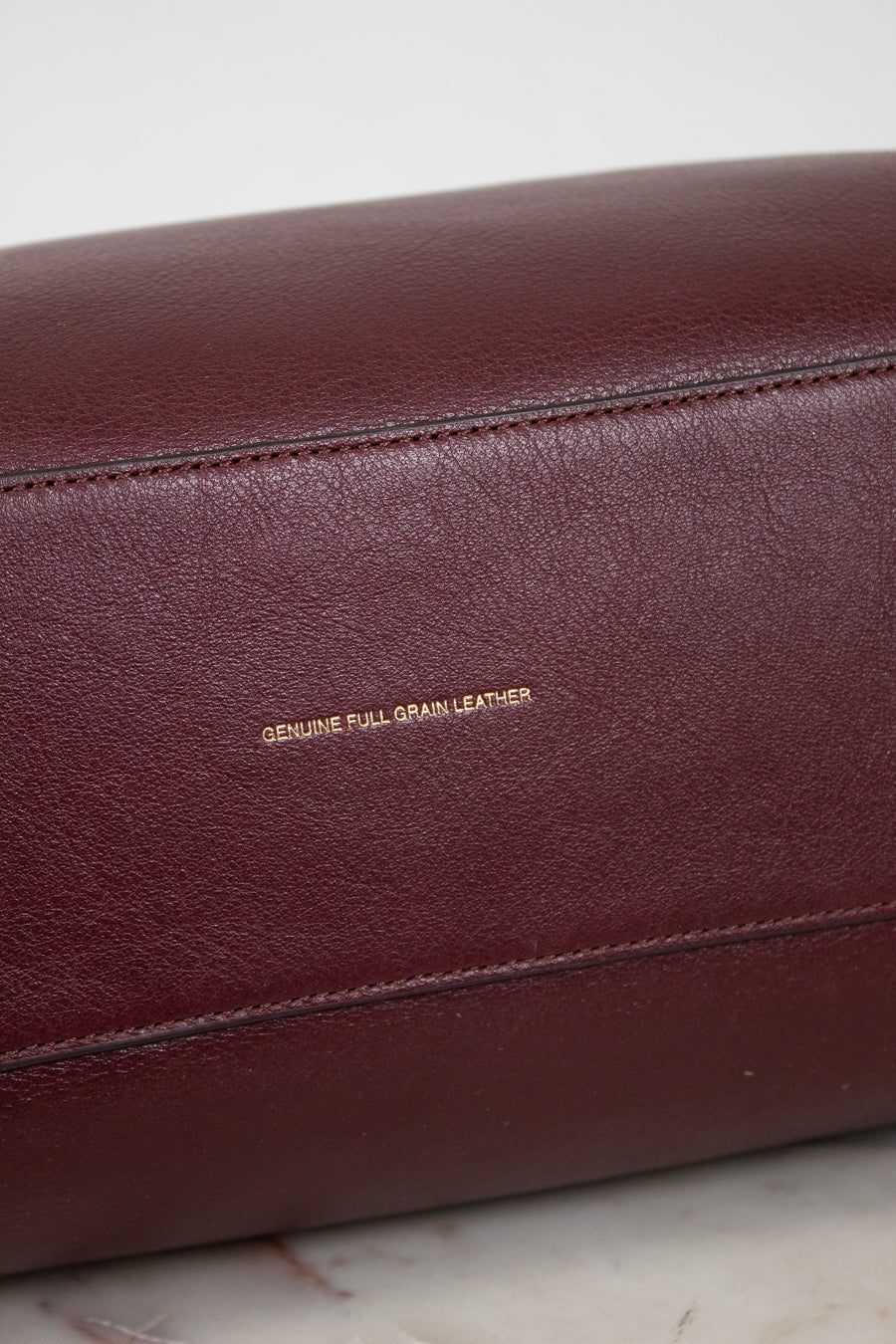 Close-up of a maroon leather bag with 'Genuine Full Grain Leather' text on a marble surface.