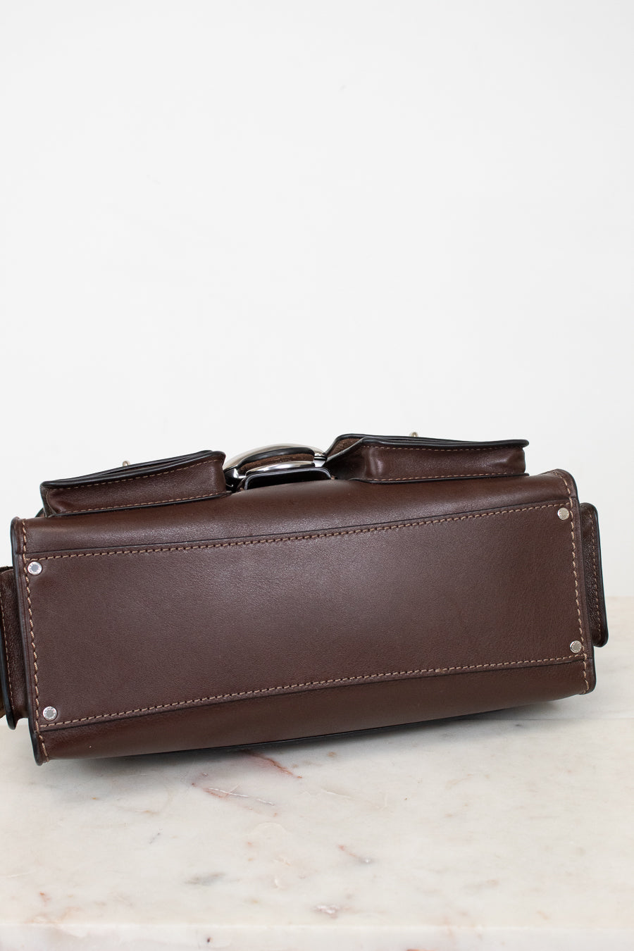 A dark brown leather tool pouch or organizer with silver metal rivets and a silver buckle, resting on a light marble surface against a white background.