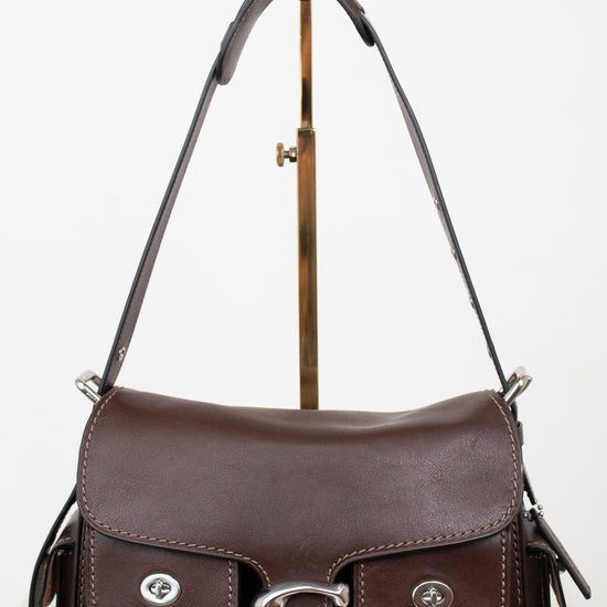 A dark brown leather shoulder bag with silver-tone hardware, displayed on a gold-colored stand against a white background.