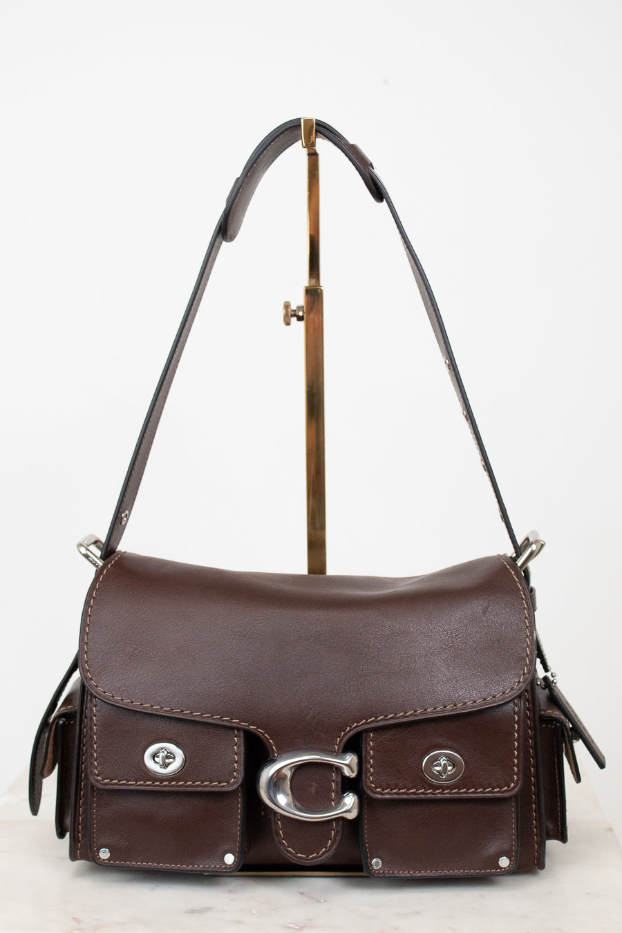 A dark brown leather shoulder bag with silver-tone hardware, displayed on a gold-colored stand against a white background.