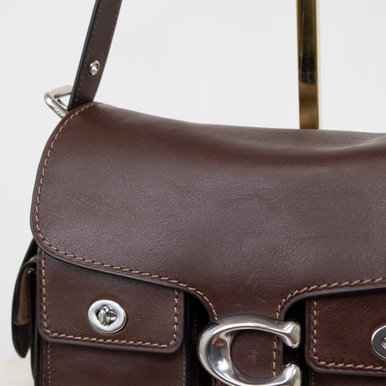 A close-up of a dark brown leather shoulder bag with silver-tone hardware, featuring a front flap pocket with a metal C-shaped buckle and a strap on a light background.