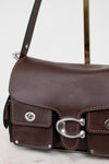 A close-up of a dark brown leather shoulder bag with silver-tone hardware, featuring a front flap pocket with a metal C-shaped buckle and a strap on a light background.