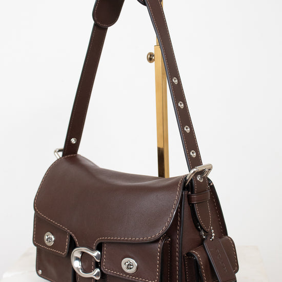 A dark brown leather shoulder bag with silver-tone hardware, displayed on a gold-colored stand against a white background.