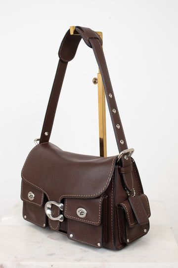 A dark brown leather shoulder bag with silver-tone hardware, displayed on a gold-colored stand against a white background.