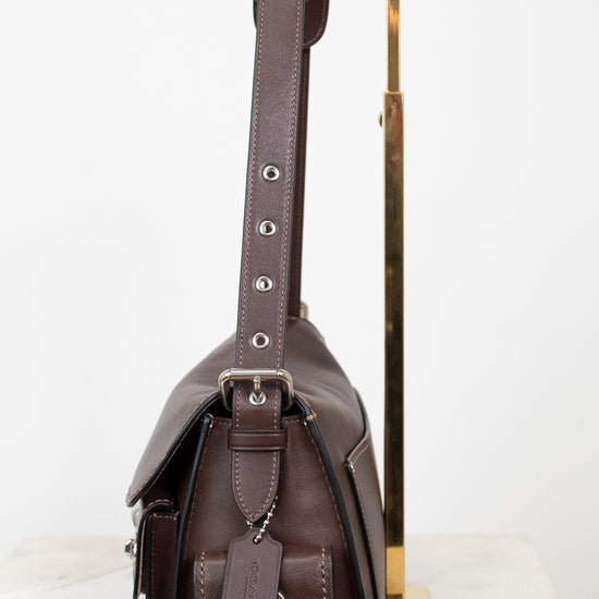 A dark brown leather shoulder bag with silver-tone hardware, shown from the side profile resting on a white marble surface next to a gold metal display stand.