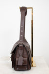 A dark brown leather shoulder bag with silver-tone hardware, shown from the side profile resting on a white marble surface next to a gold metal display stand.