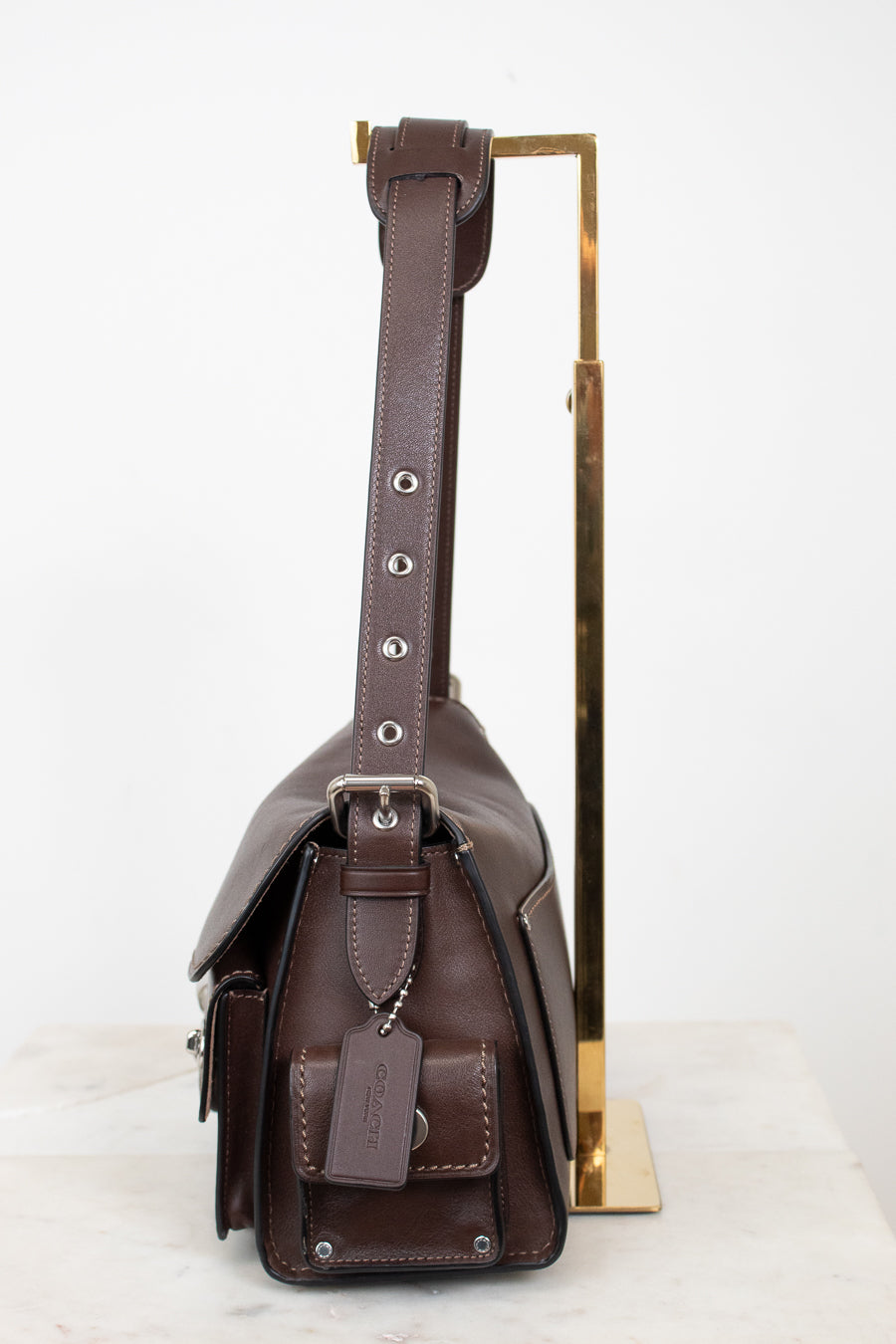 A dark brown leather shoulder bag with silver-tone hardware, shown from the side profile resting on a white marble surface next to a gold metal display stand.