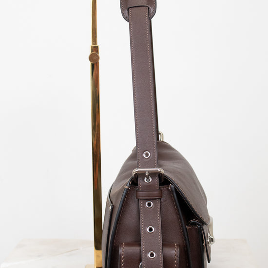 A dark brown leather shoulder bag with a wide adjustable strap, displayed on a gold-colored metal stand against a clean white background.