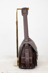 A dark brown leather shoulder bag with a wide adjustable strap, displayed on a gold-colored metal stand against a clean white background.