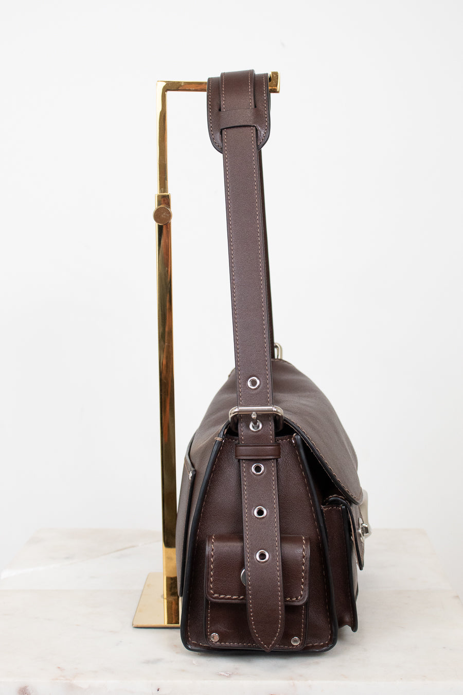 A dark brown leather shoulder bag with a wide adjustable strap, displayed on a gold-colored metal stand against a clean white background.