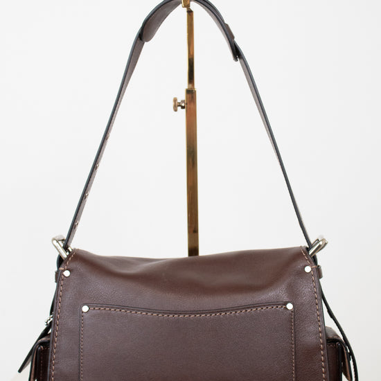 A dark brown leather shoulder bag with a front pocket and gold-tone hardware, displayed on a stand against a white background.