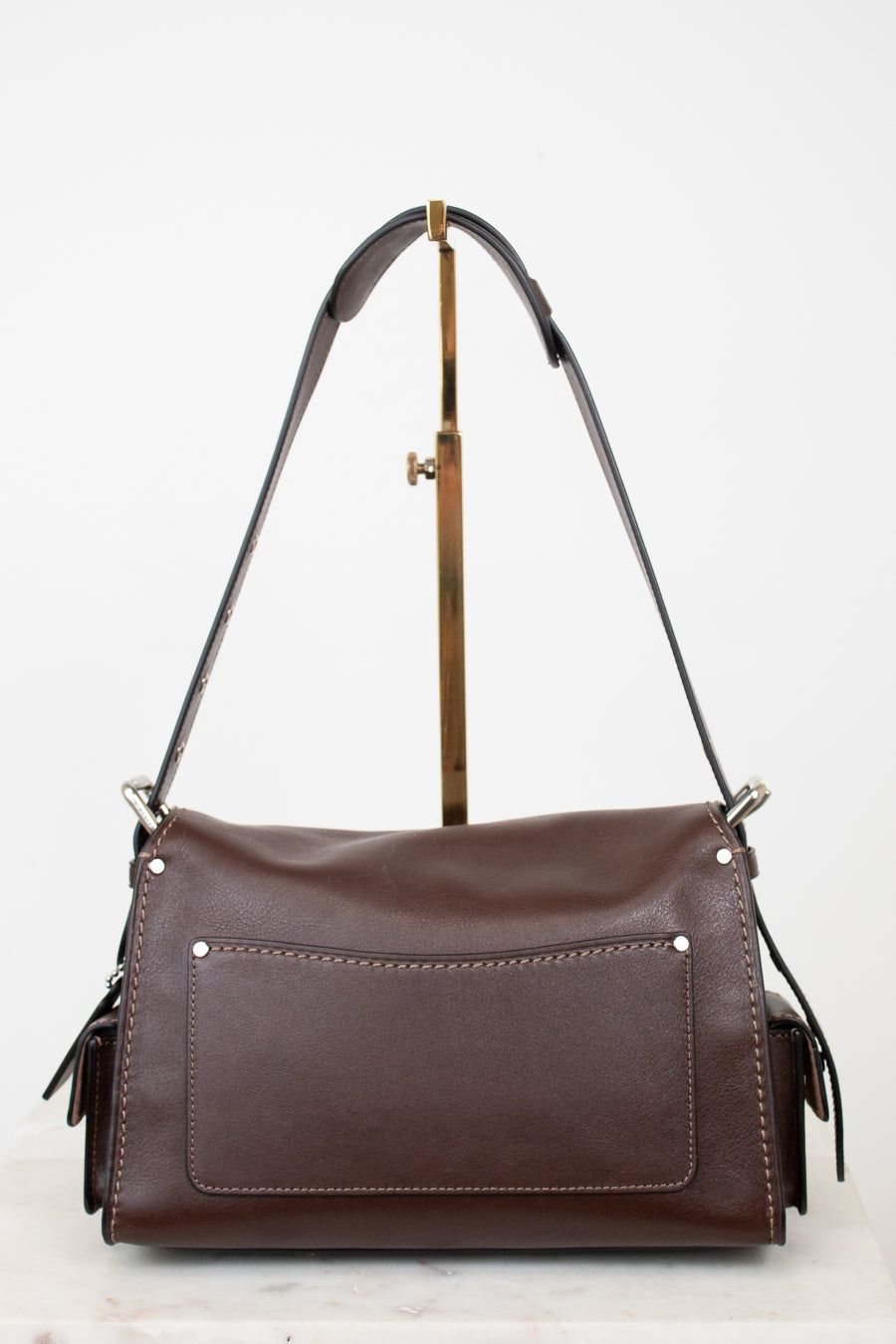 A dark brown leather shoulder bag with a front pocket and gold-tone hardware, displayed on a stand against a white background.