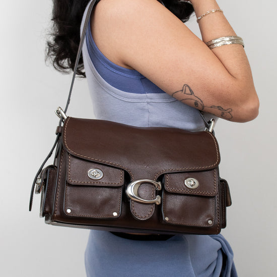A woman wearing a blue top and skirt carries a dark brown leather shoulder bag with silver-tone hardware and flap pockets against a plain light gray background.