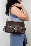 A woman wearing a blue top and skirt carries a dark brown leather shoulder bag with silver-tone hardware and flap pockets against a plain light gray background.