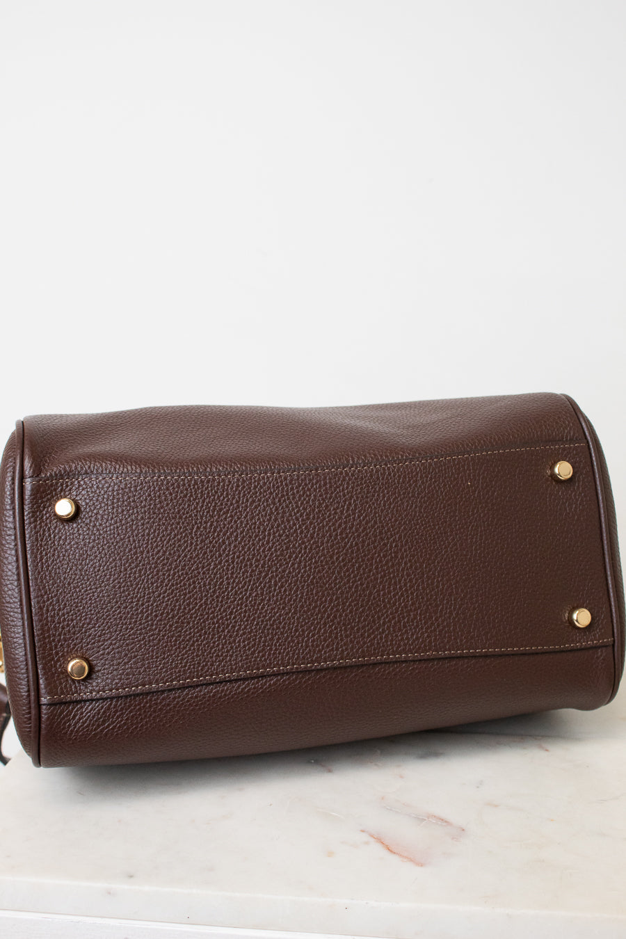 A brown pebbled leather handbag shown from the bottom, resting on a white marble surface against a white background.