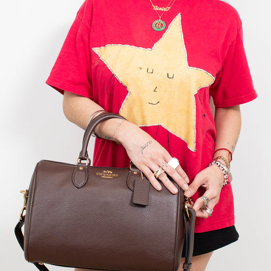 A model wearing a red graphic t-shirt and black shorts holds a brown pebbled leather top-handle handbag with a gold logo. The background is a clean white studio wall.