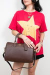A model wearing a red graphic t-shirt and black shorts holds a brown pebbled leather top-handle handbag with a gold logo. The background is a clean white studio wall.