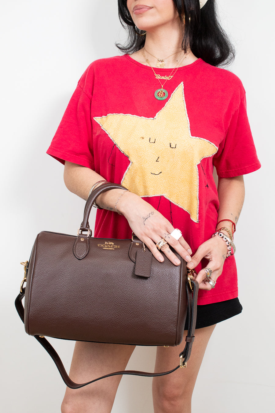 A model wearing a red graphic t-shirt and black shorts holds a brown pebbled leather top-handle handbag with a gold logo. The background is a clean white studio wall.