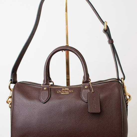 A dark brown pebbled leather Coach satchel with gold-tone hardware, displayed on a white background with a gold stand.