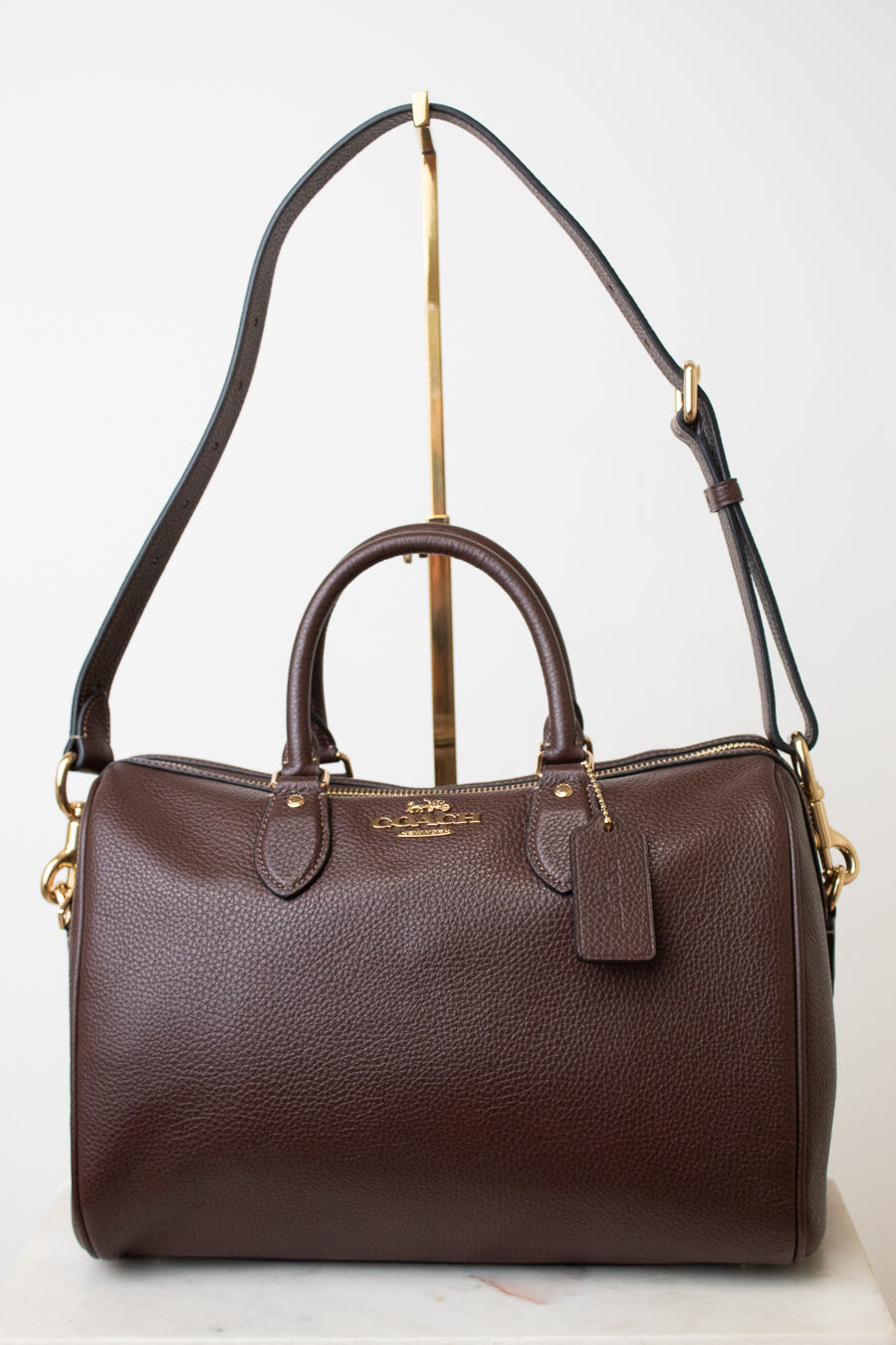 A dark brown pebbled leather Coach satchel with gold-tone hardware, displayed on a white background with a gold stand.