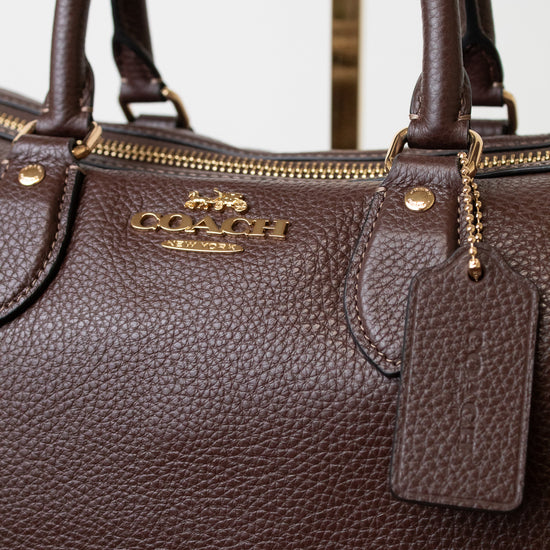 Close-up of a dark brown pebbled leather Coach handbag with gold-tone hardware, a top zipper, and a hanging leather tag. The Coach logo is visible on the front.