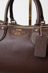 Close-up of a dark brown pebbled leather Coach handbag with gold-tone hardware, a top zipper, and a hanging leather tag. The Coach logo is visible on the front.