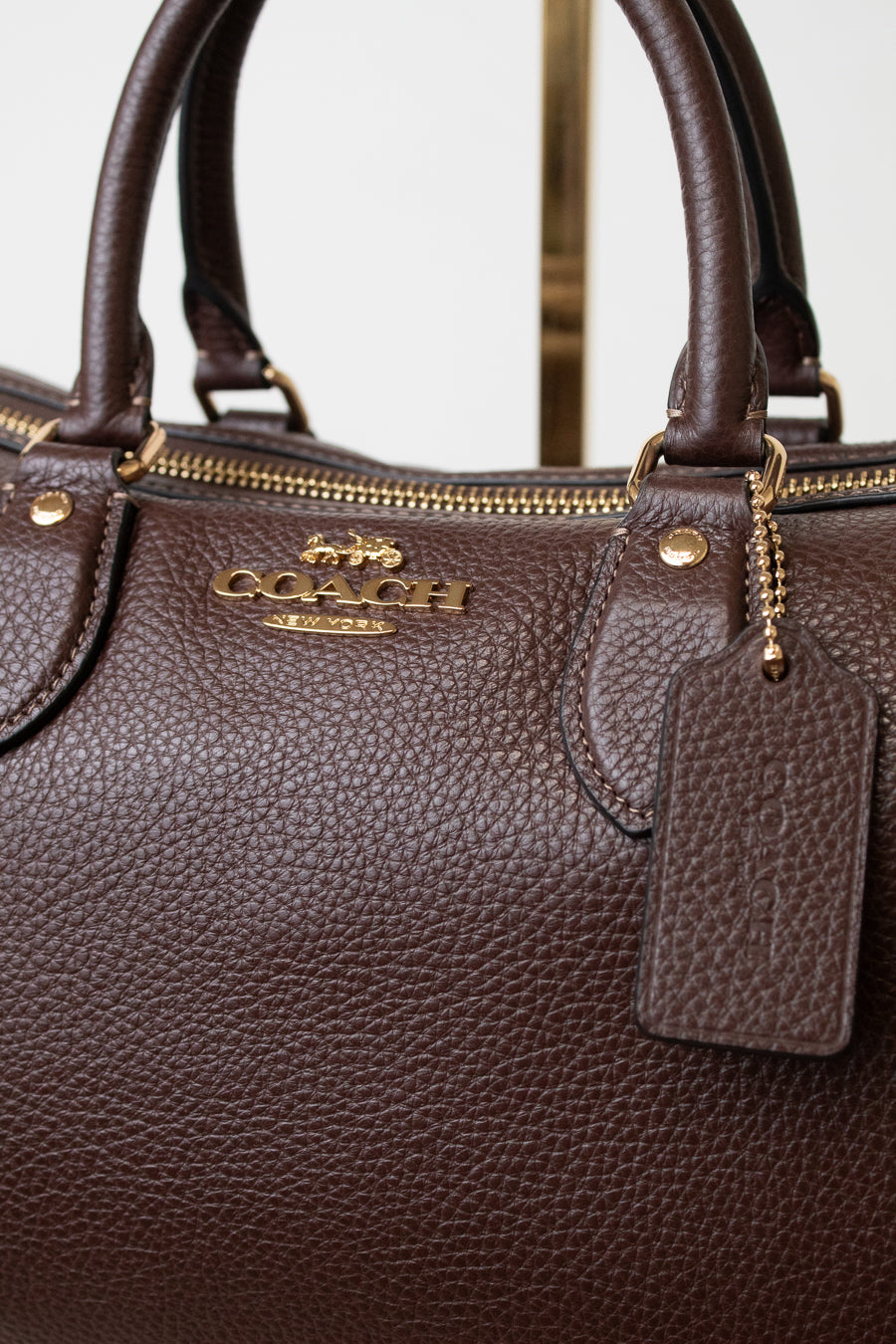 Close-up of a dark brown pebbled leather Coach handbag with gold-tone hardware, a top zipper, and a hanging leather tag. The Coach logo is visible on the front.