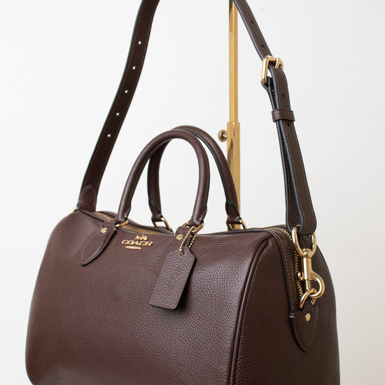 A dark brown pebbled leather Coach satchel with gold-tone hardware, displayed on a white marble surface against a white background.