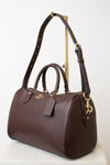 A dark brown pebbled leather Coach satchel with gold-tone hardware, displayed on a white marble surface against a white background.
