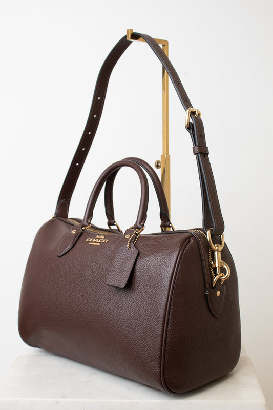A dark brown pebbled leather Coach satchel with gold-tone hardware, displayed on a white marble surface against a white background.