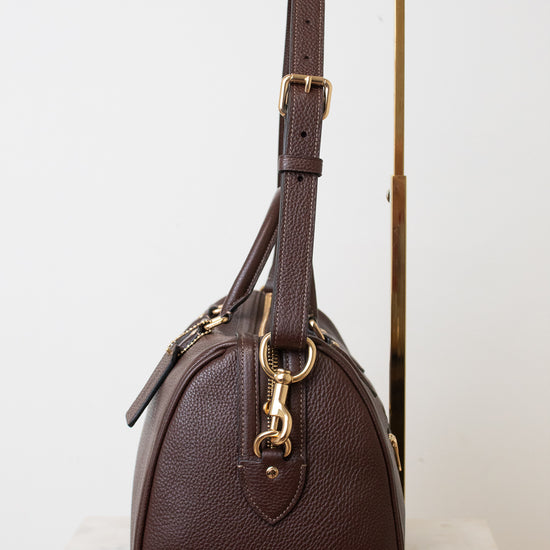 A dark brown pebbled leather handbag with gold-tone hardware, displayed on a minimalist gold stand against a clean white background.