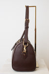 A dark brown pebbled leather handbag with gold-tone hardware, displayed on a minimalist gold stand against a clean white background.