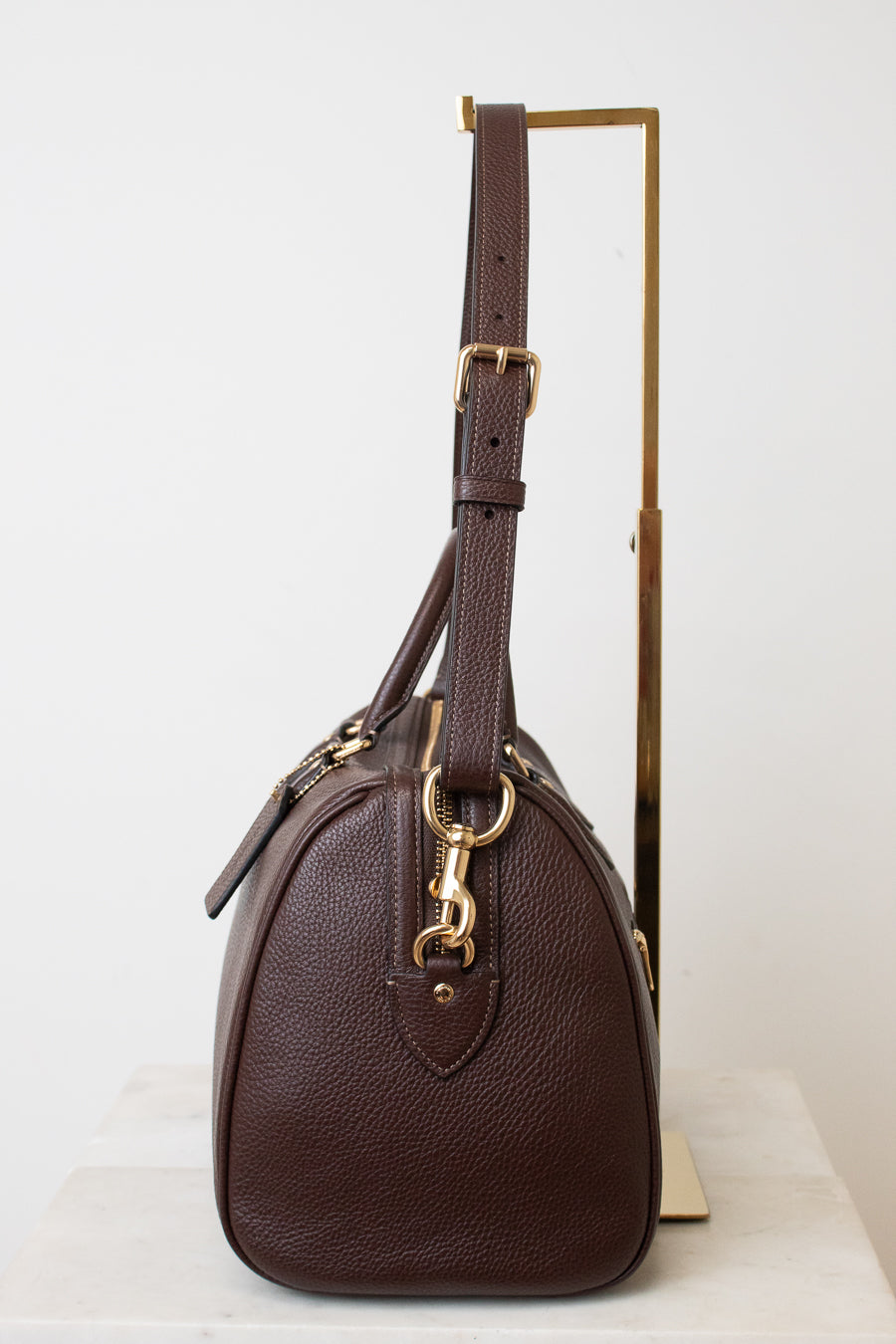 A dark brown pebbled leather handbag with gold-tone hardware, displayed on a minimalist gold stand against a clean white background.