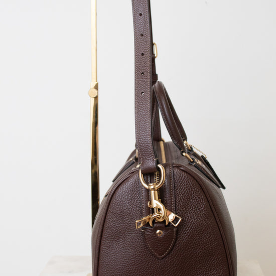 A dark brown pebbled leather handbag with gold-tone hardware, displayed on a minimalist gold metal stand against a clean white background.