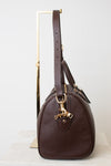 A dark brown pebbled leather handbag with gold-tone hardware, displayed on a minimalist gold metal stand against a clean white background.
