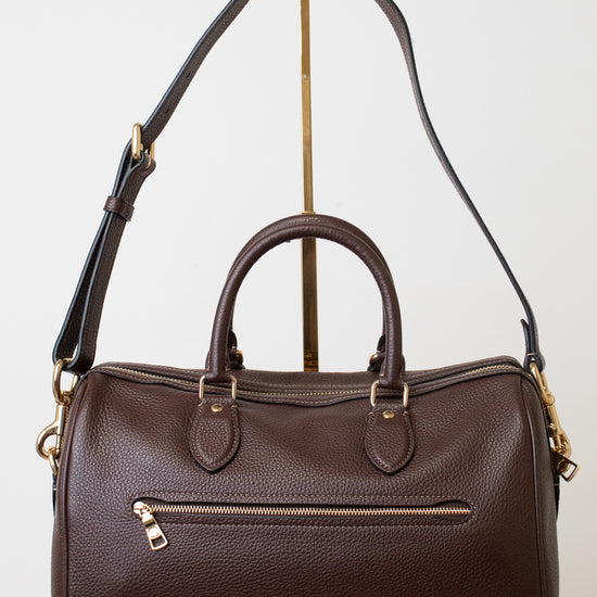 A dark brown pebbled leather satchel with double top handles, an exterior zippered pocket, and adjustable shoulder strap, displayed on a gold-colored stand against a white background.