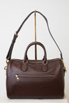 A dark brown pebbled leather satchel with double top handles, an exterior zippered pocket, and adjustable shoulder strap, displayed on a gold-colored stand against a white background.