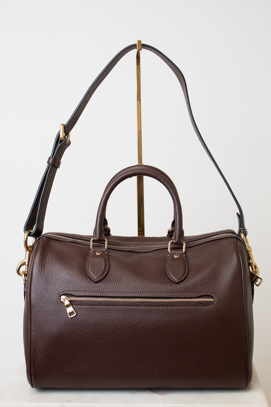 A dark brown pebbled leather satchel with double top handles, an exterior zippered pocket, and adjustable shoulder strap, displayed on a gold-colored stand against a white background.