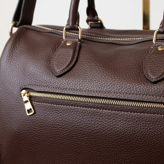 A close-up of a dark brown pebbled leather handbag with gold-tone hardware, including an exterior zip pocket and top handles, set against a light neutral background.