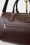 A close-up of a dark brown pebbled leather handbag with gold-tone hardware, including an exterior zip pocket and top handles, set against a light neutral background.