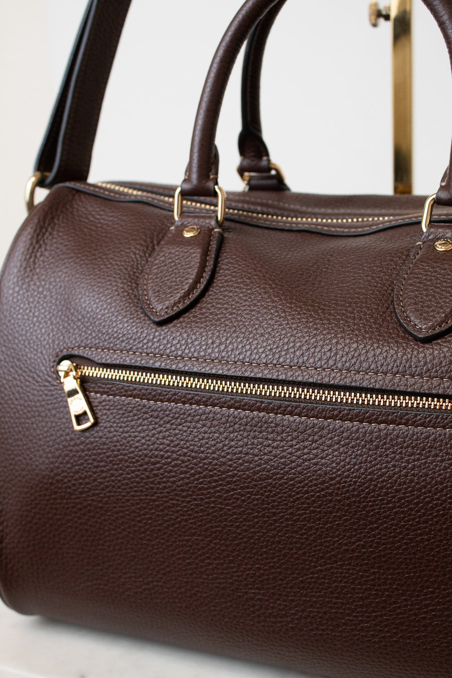 A close-up of a dark brown pebbled leather handbag with gold-tone hardware, including an exterior zip pocket and top handles, set against a light neutral background.