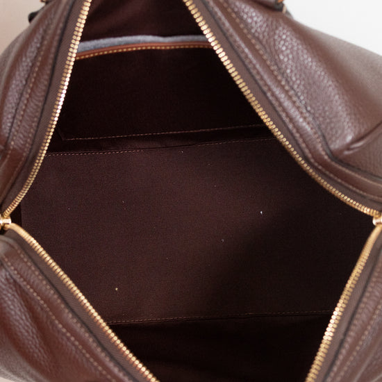 A close-up of a brown pebbled leather handbag being held open by a hand, showing the interior lining and gold-tone zipper.
