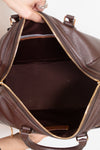 A close-up of a brown pebbled leather handbag being held open by a hand, showing the interior lining and gold-tone zipper.