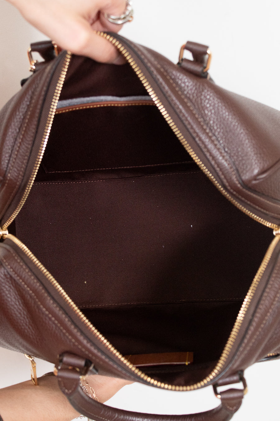 A close-up of a brown pebbled leather handbag being held open by a hand, showing the interior lining and gold-tone zipper.
