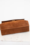 A brown suede clutch bag with a gold-tone metal clasp, resting on a white marble surface against a white background.