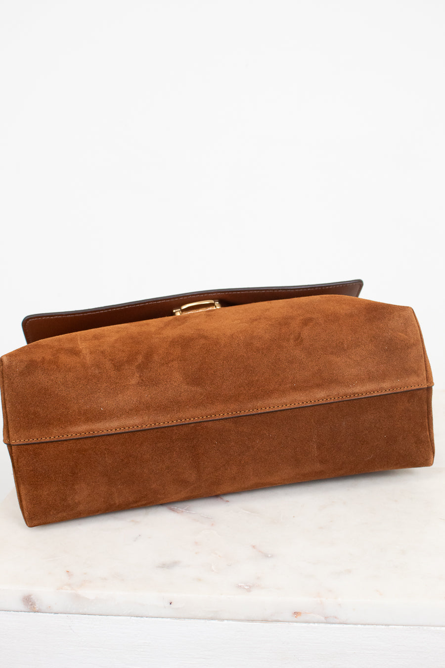 A brown suede clutch bag with a gold-tone metal clasp, resting on a white marble surface against a white background.